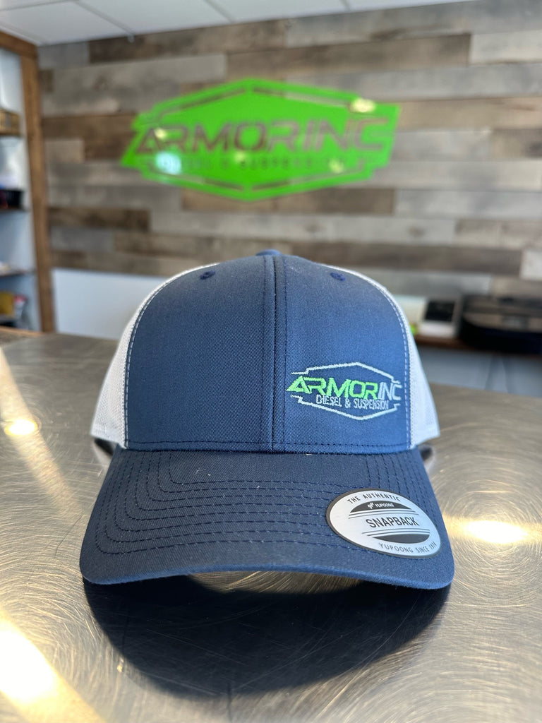 A navy blue and gray Adjustable Trucker Armor Inc Hat with the Armor Inc Diesel & Suspension logo embroidered on the front, displayed on a metal counter with a green neon sign in the background.